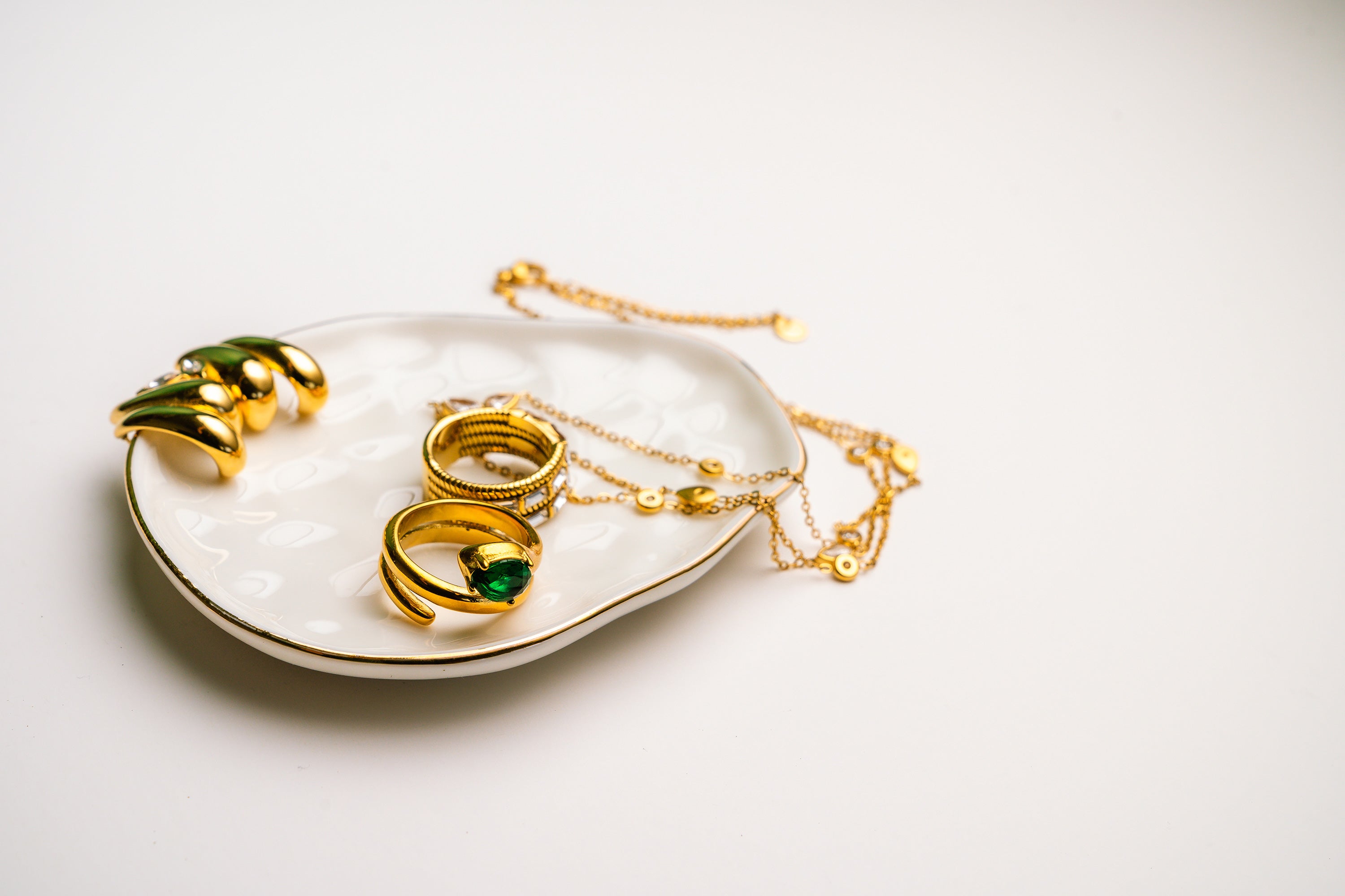 Gold jewelry including rings and a necklace on a white dish with a white background