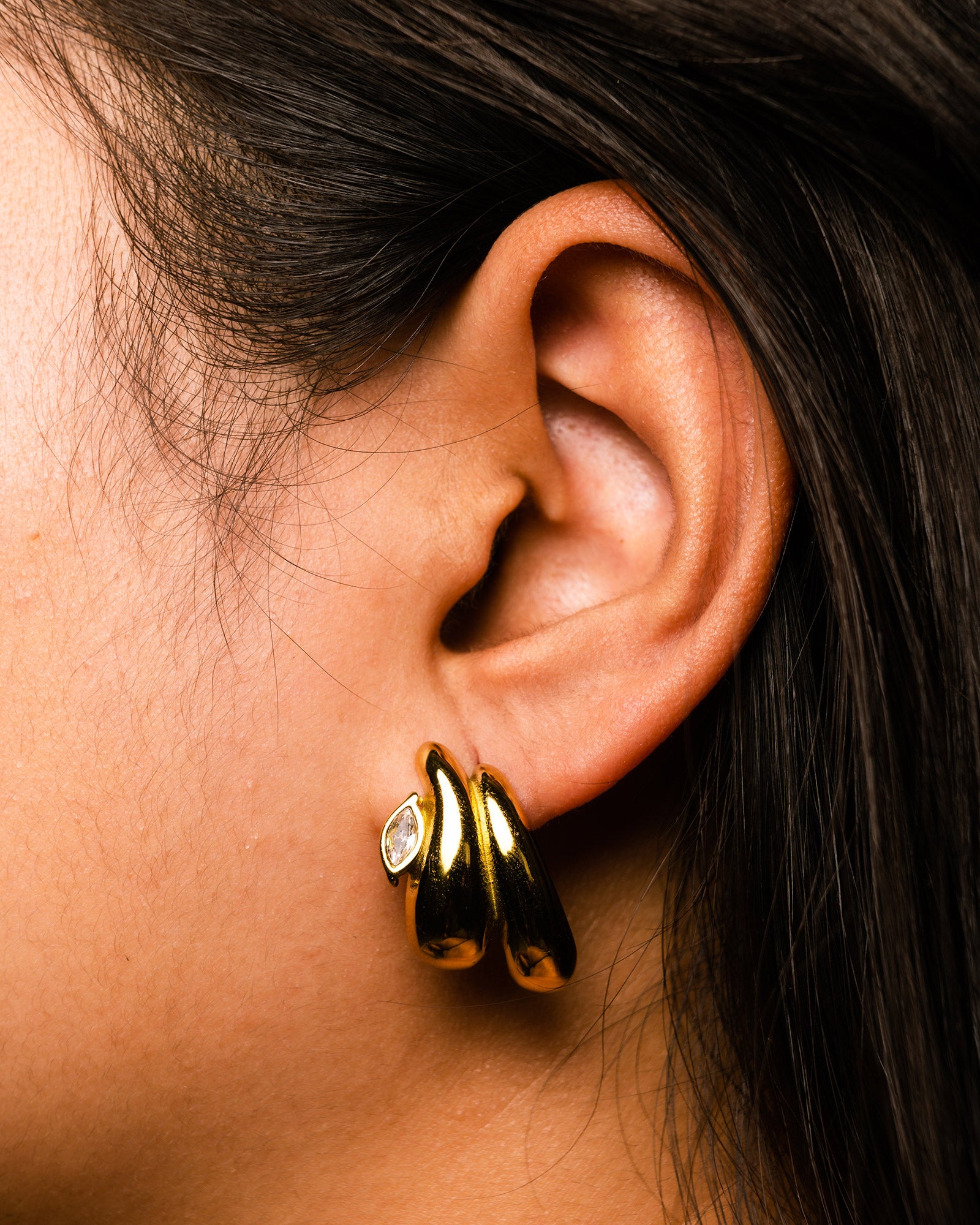 Close-up of an ear wearing a gold double teardrop golden studs earring with a clear gemstone.