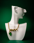 Gold midnight muse necklace with a green heart pendant on a white mannequin bust against a green background