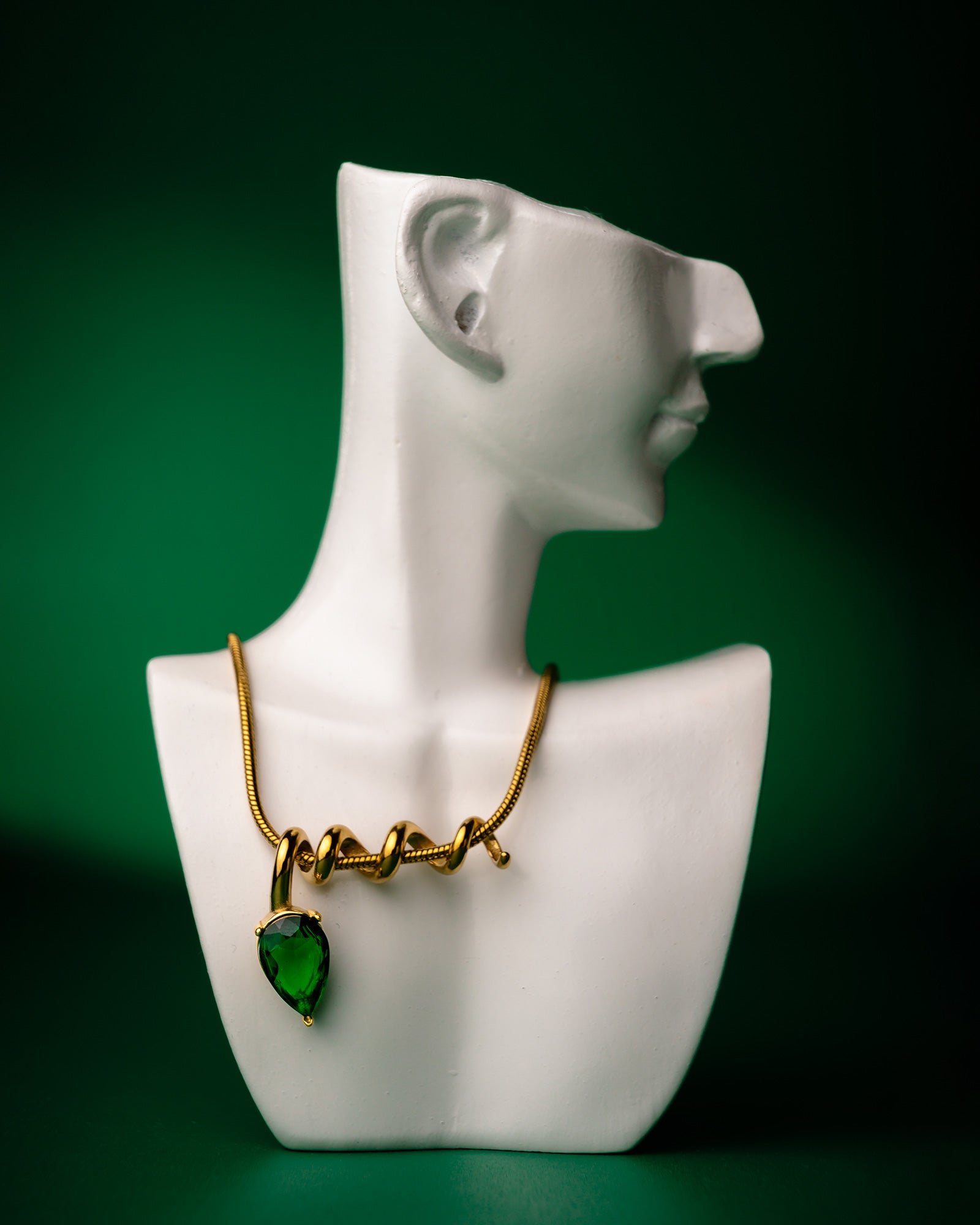 Gold midnight muse necklace with a green heart pendant on a white mannequin bust against a green background