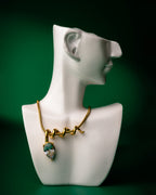 Gold midnight muse necklace with a green pendant on a white ceramic bust against a dark green background